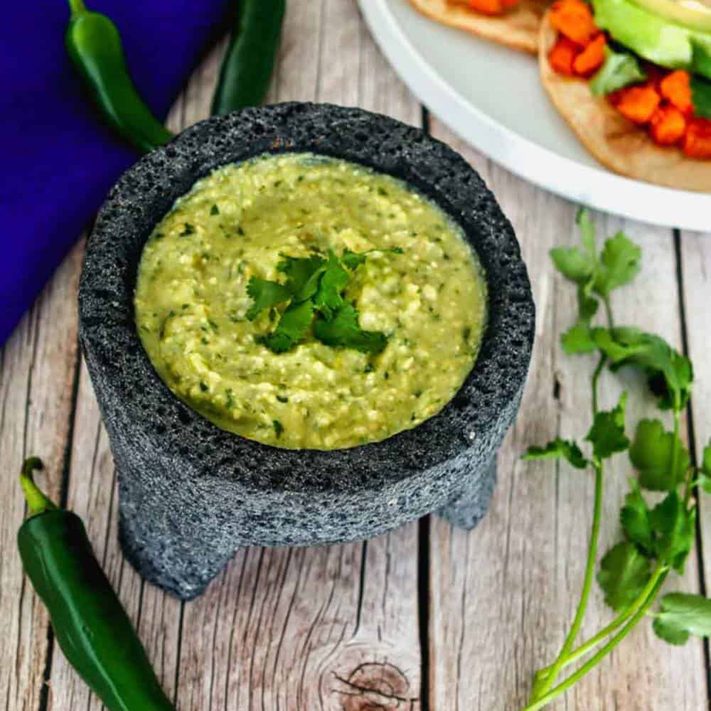 Salsa verde in a molcajete. Some chiles and cilantro around the molcajete. Two sweet potato tacos in the top right corner.