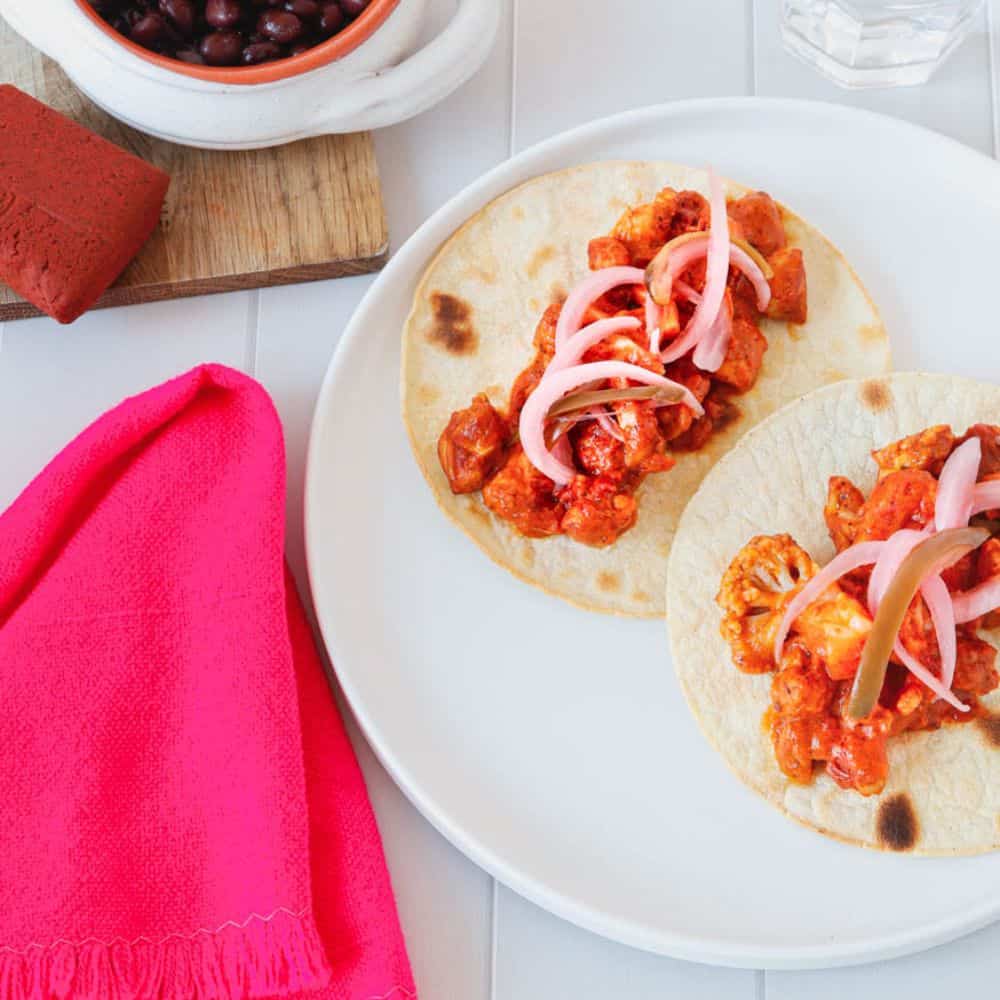 Achiote chicken tacos in a plate. Pink table cloth on the side and black beans in a white bowl on the top left corner.
