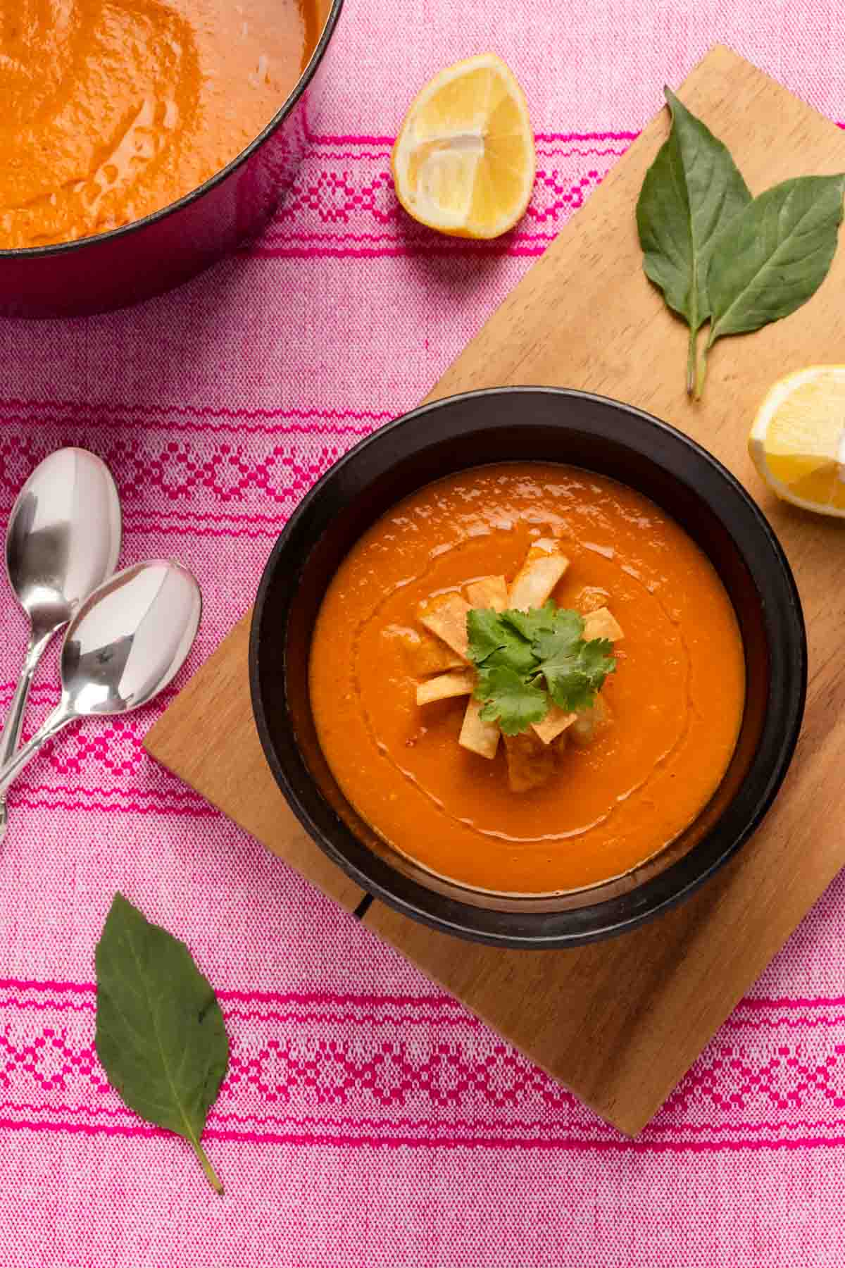 Carrot Tomato Soup served in a brown bowl with tortilla strips and chopped cilantro on top.