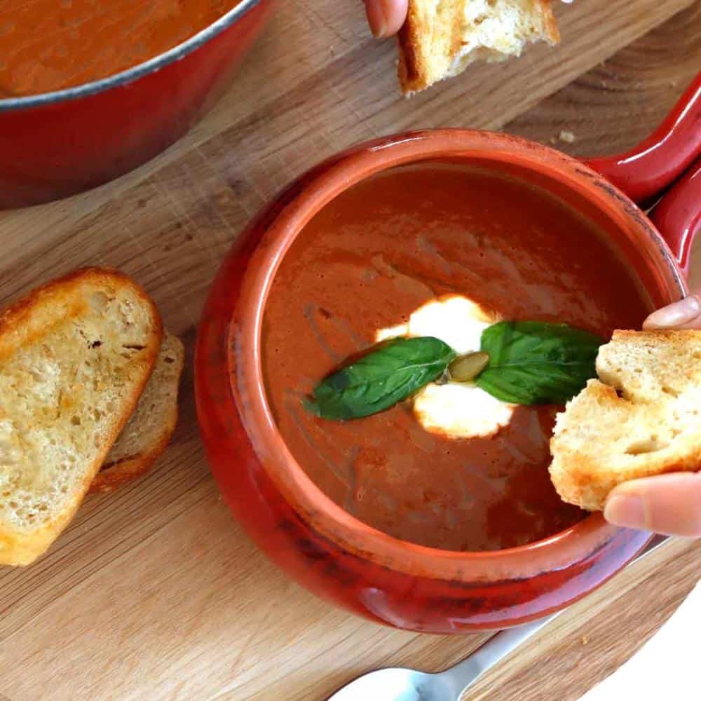 A bowl of tomato soup and someone ready to eat it with some pieces of sourdough bread.