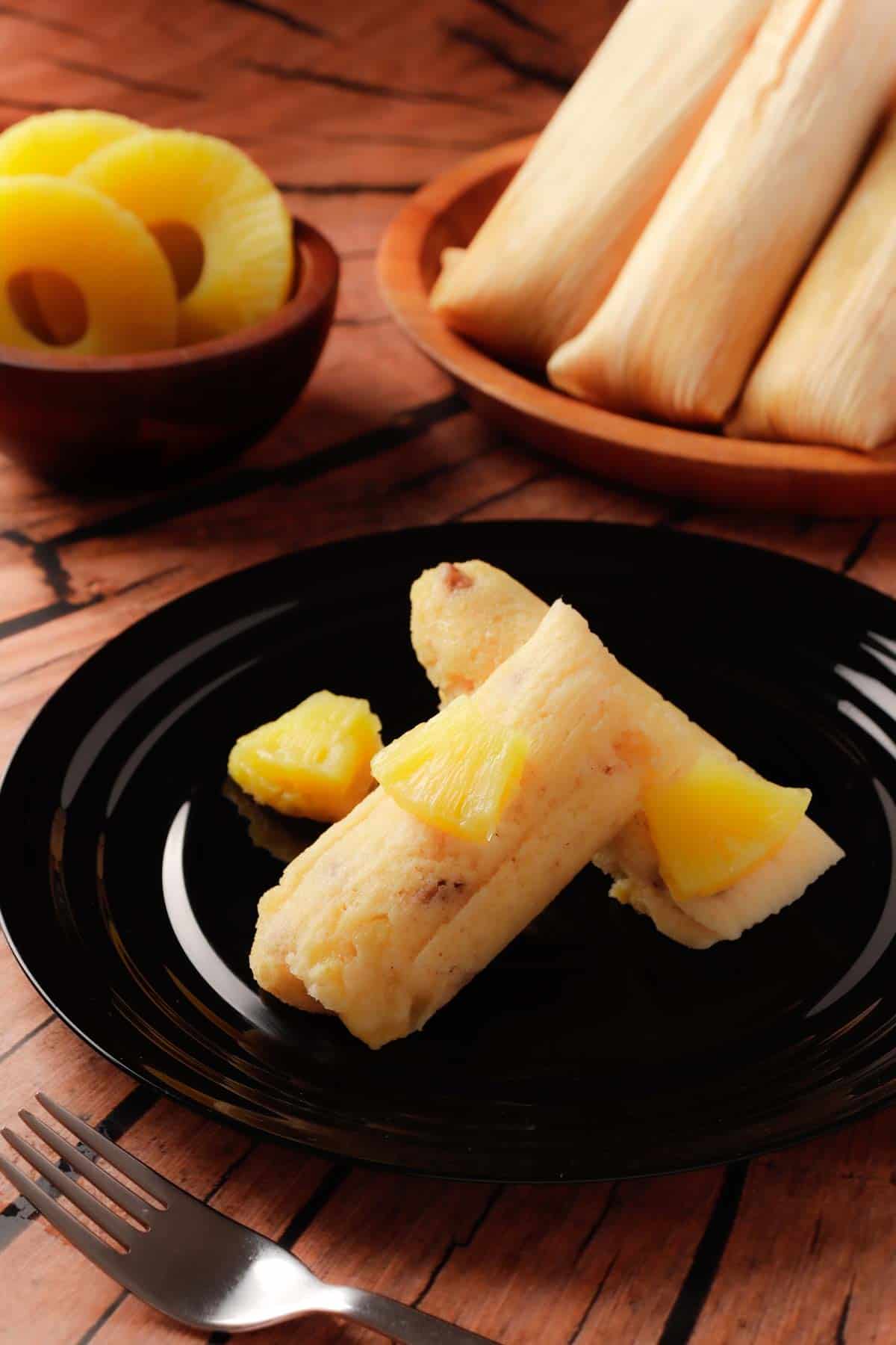 Two sweet pineapple Mexican tamales in a black plate and some more pineapple tamales in the background.