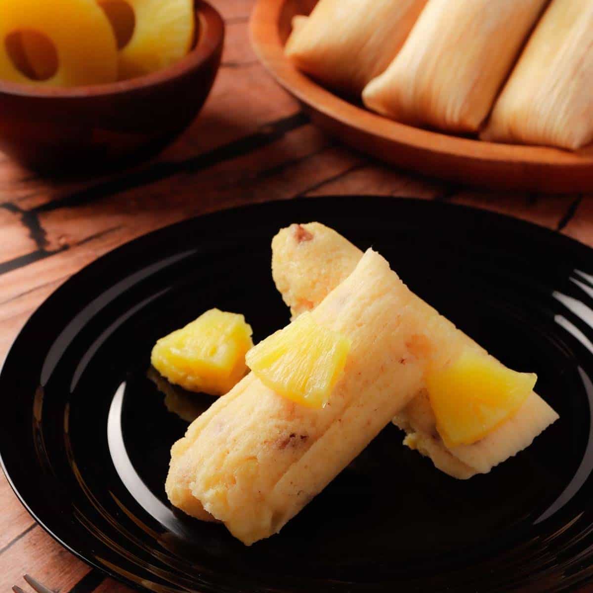 Two sweet pineapple Mexican tamales in a black plate and some more pineapple tamales in the background.