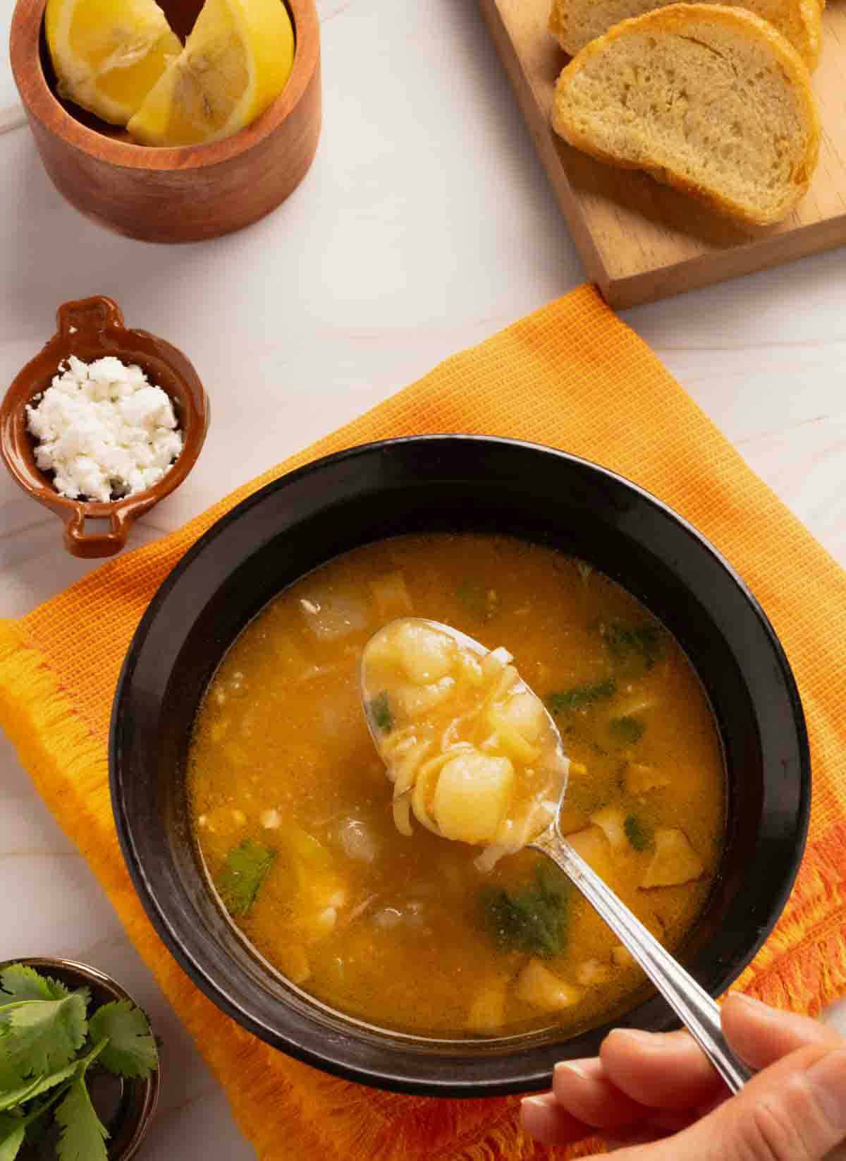Potato and leek soup served in a bowl surrounded by lemon wedges, queso fresco and bread slices.