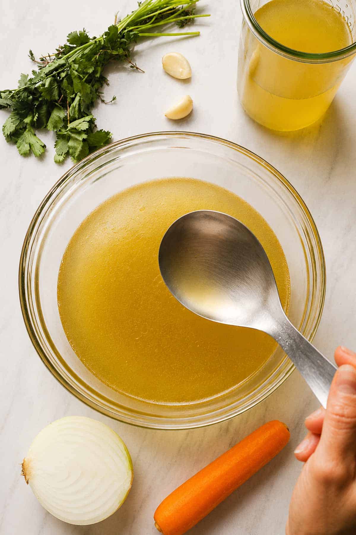 Chicken broth served in a transparent bowl and some of the ingredients to make it are in the picture.